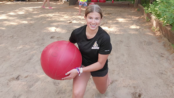 An image of a girl doing lunges in the sand holding a large red water ball