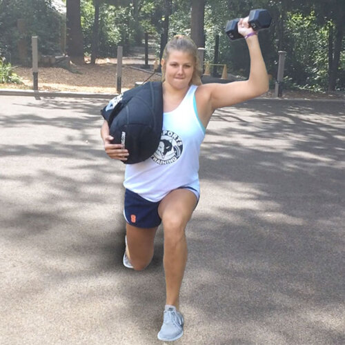 An image of a girl doing a lunge while holding a weighted dumbbell in one hand and holding a sandbag on her shoulder