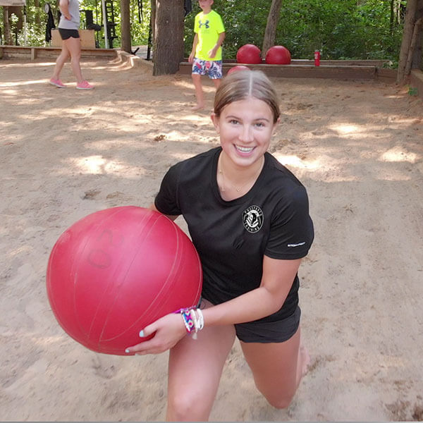 An image of a girl doing lunges in the sand holding a large red water ball